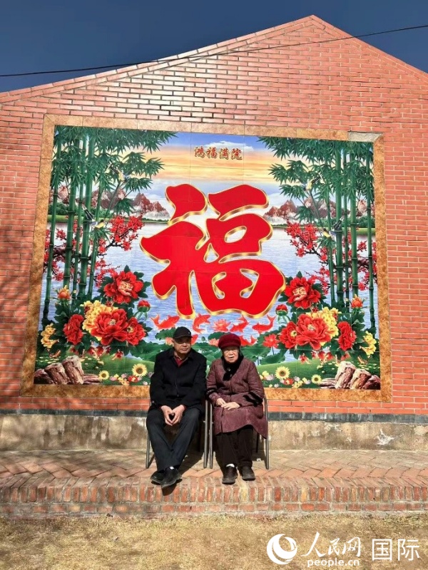 在莫尼山非遗小镇的“福”字墙前，晒着太阳休息的一对老夫妻。人民网 河贞美摄