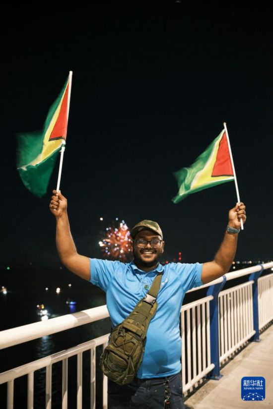10月5日,在圭亚那首都乔治敦,一名男子挥舞圭亚那国旗在巴拉特·贾格迪奥-德梅拉拉河大桥上与身后的烟花合影。新华社记者 许咏政 摄